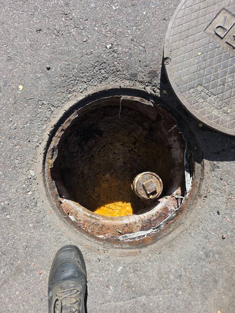 Open grease interceptor manhole in an asphalt lot, with its lid set beside it and a crew member's boot in the foreground